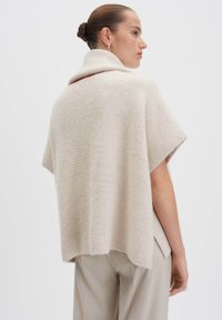 Donna con i capelli raccolti in un bun, che indossa un maglione a collo alto a coste beige a maniche corte e pantaloni beige chiaro, in posizione laterale su uno sfondo bianco.