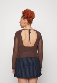 Top castanho de mangas longas com costas abertas, apresentando um fecho com laço. Combinado com uma saia curta mini com riscas azuis. Textura suave, design casual.