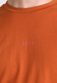 Kortermet oransje t-skjorte med rund hals og en hevet rosa "BOSS"-logo på brystet, laget av myk bomullsmateriale.