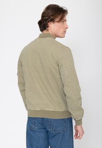 Man met golvend bruin haar, gekleed in een licht olijfgroene bomberjas en blauwe spijkerbroek, met zijn rug naar de camera en zijwaarts kijkend tegen een effen achtergrond.