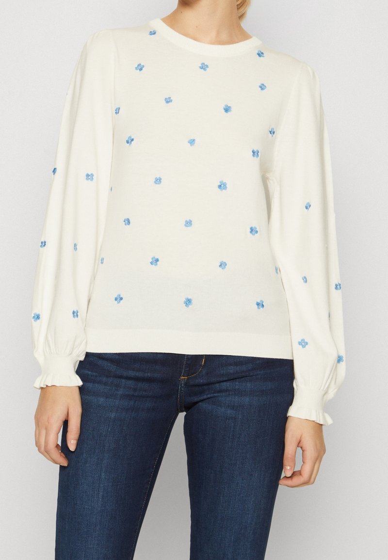 Vrouw in een witte trui met lange mouwen en kleine blauwe bloemmotieven, met een donkerblauwe jeans, staande tegen een effen lichte achtergrond.