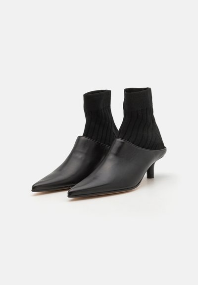 Melnas ādas kurpes ar smailiem pirkstiem un bloku papēdi, ar ribotu melnu adītu augšdaļu, kas izskatās kā zeķes. Gluda tekstūra ar minimālistisku dizainu.