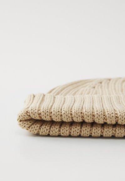Chapeau en tricot beige, présentant une texture côtelée et un revers replié. La laine douce avec des détails de couture visibles ajoute de la dimension.