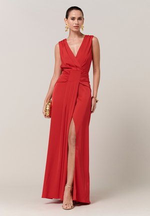 Žena v červenom šatách bez rukávov s hlbokým výstrihom do V a rozparkom nad stehno, doplnená zlatými náušnicami, náramkom, psaníčkom a sandálmi na remienky.