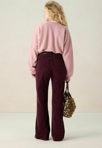 Femme aux longs cheveux blonds, portant un pull rose et un pantalon violet foncé, tenant un sac à main en peluche à imprimé léopard, de dos face à la caméra.
