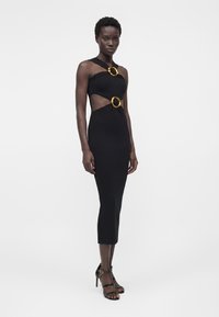 Rochie midi neagră mulată, cu decupaje și două catarame aurii mari, rotunde, purtată cu sandale negre cu toc înalt pe un model în picioare.
