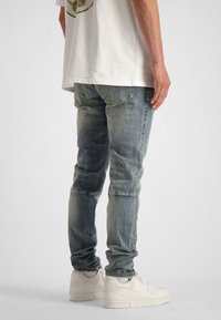 Smala blå denimjeans med blekta områden, sömnadsdetaljer och en sliten textur, i kombination med vita sneakers och en enkel vit T-shirt.