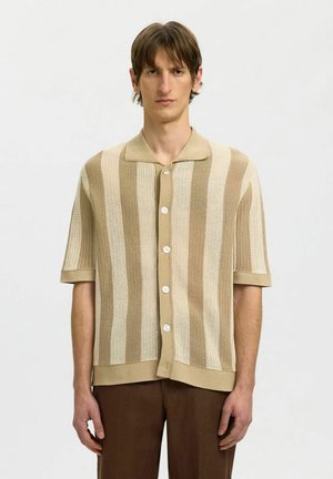 Mężczyzna ubrany w beżowo-kremowy, pionowo prążkowany, krótki sweter zapinany na guziki, w połączeniu z brązowymi spodniami, stojący na jednolitym tle.