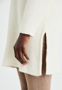 Pull en tricot côtelé ivoire avec des manches longues et des fentes sur les côtés. La texture est douce et duveteuse, avec un ourlet légèrement effiloché.