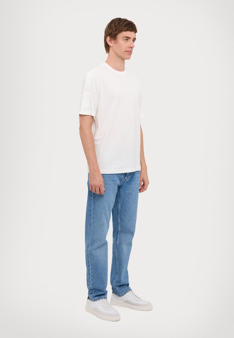 Bílá bavlněná T-shirt s krátkým rukávem, kombinovaná s světle modrými džíny a bílými teniskami, stojící na jednolitém pozadí.