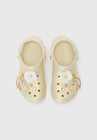 Gold Crocs with a textured finish, featuring a white flower and decorative jewels. Includes a strap and several cut-out holes for ventilation.