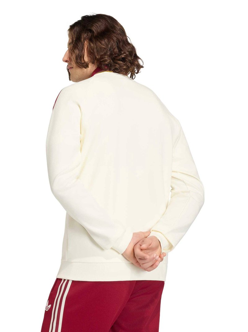 Personne aux cheveux bouclés, vue de profil vers la gauche, portant un haut blanc à manches longues et un pantalon de survêtement rouge Adidas avec des bandes blanches.