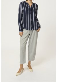 Frau trägt eine marineblaue und weiße vertikal gestreifte Bluse, hellgraue, verkürzte Hose und beige, flache Slingback-Schuhe und steht mit den Händen in den Taschen.