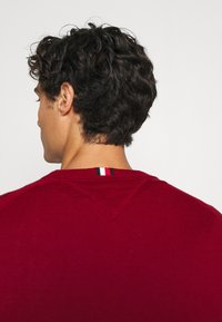 Maglione rosso lavorato a maglia con collo tondo, caratterizzato da un dettaglio a punto V sul retro e una piccola striscia tri-color al collo.