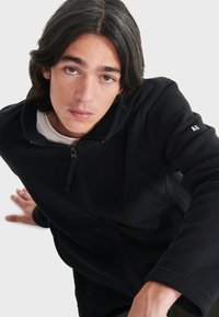 Schwarze Fleece-Zipjacke mit hohem Kragen und elastischen Bündchen. Verfügt über einen Frontreißverschluss und ein kleines Logo-Detail am Ärmel.