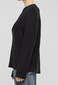 Kobieta w czarnym swetrze o prążkowanej strukturze z długimi rękawami, w połączeniu z jasnoniebieskimi dżinsami, pokazana z boku na jednolitym tle.