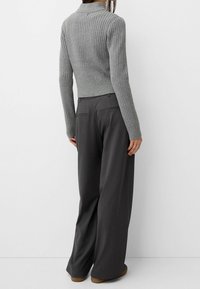 Kobieta stoi tyłem, ubrana w dopasowany szary prążkowany sweter i luźne, ciemnoszare spodnie o szerokich nogawkach oraz brązowe buty.