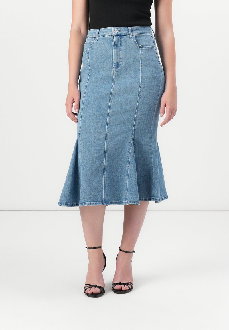 Vrouw in een aansluitend zwart top en een lange spijkerrok, staand op stijlvolle zwarte sandalen met bandjes, tegen een eenvoudige witte achtergrond.