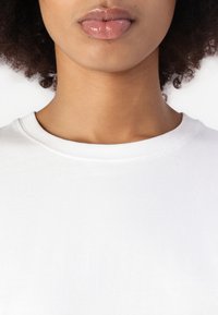 T-shirt bianco in cotone con scollo rotondo, caratterizzato da una texture liscia e un sottile dettaglio di cucitura; parte superiore del corpo del modello visibile, rivolto in avanti.
