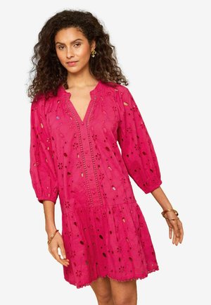 Frau mit lockigem Haar trägt ein hellpinkes, mit Blumenmuster durchbrochenes Kleid mit Dreiviertelärmeln und goldenen Creolen und posiert vor einem einfarbigen Hintergrund.