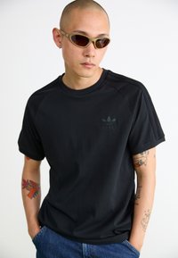 Czarna bawełniana koszulka z teksturowanym logo adidas, krótki rękaw i fason raglanowy; w komplecie z okrągłymi okularami przeciwsłonecznymi i niebieskimi dżinsami.
