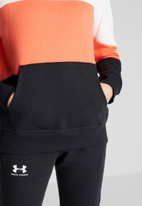 Sudadera color bloque rojo y blanco con un bolsillo tipo canguro y puños de canalé, combinada con joggers negros que tienen un logo blanco.