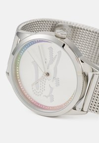 Reloj con correa de malla plateada y esfera blanca, diseño metálico de cocodrilo y bisel de cristales multicolores. Caja redonda y marcadores de hora simples.
