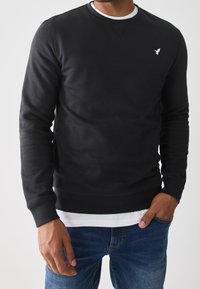 Hombre vistiendo un sudadera negra de cuello redondo sobre una camiseta blanca y jeans azules, con la mano en el bolsillo, sobre un fondo claro sencillo.