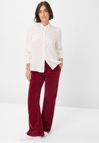 Witte overhemd met knopen en kraag, gecombineerd met bordeauxrode corduroy flare broeken. Model draagt witte sneakers en staat tegen een neutrale achtergrond.