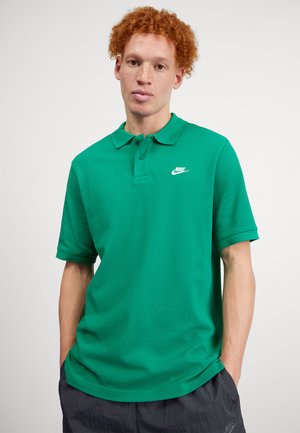Camisa polo verde de algodón con cuello clásico, mangas cortas y un logo blanco de Nike en el lado izquierdo del pecho. Textura suave.