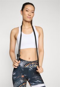 Biely športový top a čierne legíny s farebným, abstraktným vzorom. Modelka drží čierny skákač, ktorý má textúrovanú rukoväť.