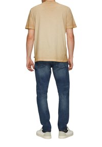 Beige T-Shirt mit kurzen Ärmeln und leicht verblasstem Finish, kombiniert mit blauen Jeans. Das Outfit zeichnet sich durch eine lockere Passform und ein simples Design aus. Weiße Sneakers.