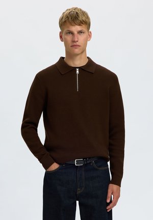 Homme blond portant un pull en maille marron foncé à manches longues avec un col zippé et un jean bleu foncé, debout avec une main dans la poche.