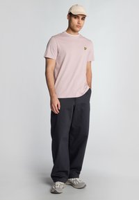 Lys pink kortærmet t-shirt med et lille logo, parret med mørke løse bukser og lyse grå sneakers, toppet med en beige kasket.