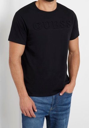 Mężczyzna ubrany w czarny T-shirt z krótkim rękawem i wypukłym logo "GUESS" na klatce piersiowej oraz niebieskie dżinsy, ręka w kieszeni, stoi na tle białego tła.