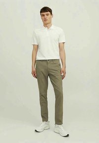 Hvid poloshirt, lysegrå bukser og hvide sneakers. Bukserne er lige skåret med forlommer og bæltestropper. Ensfarvet baggrund.