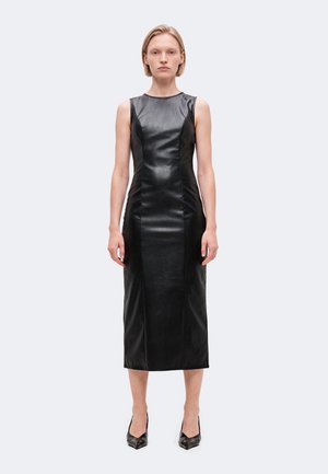 Le mannequin se tient de face, vêtue d’une robe midi en cuir noir sans manches et de chaussures à talons noires, sur un fond uni et clair.