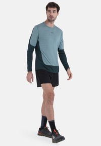Langärmliges, leichtes Sportshirt in Türkis und Dunkelgrün, kombiniert mit schwarzen Shorts und grauen Laufschuhen mit orangefarbenen Akzenten.