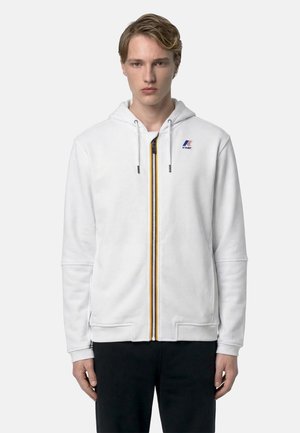 Giovane uomo che indossa una felpa bianca con cappuccio, cerniera frontale e logo K-Way, in piedi davanti a uno sfondo chiaro e uniforme.