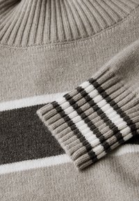 Beige neule, jossa on korkea ribattu kaulus, ja tummanharmaa sekä valkoinen raidallinen kuvio keholla sekä ribattu hihansuu, jossa on tummia raitoja.
