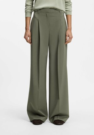 Personne portant un pantalon taille haute, jambes larges, vert olive avec plis, associé à des chaussures marron foncé, debout devant un fond uni.