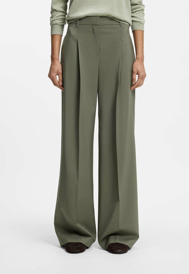 Personne portant un pantalon taille haute, jambes larges, vert olive avec plis, associé à des chaussures marron foncé, debout devant un fond uni.
