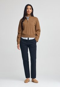 Brauner Zip-Hals-Pullover aus weichem Stoff, der über einem hellblauen Kragenhemd getragen wird, kombiniert mit schwarzen Hosen und braunen Slippern.