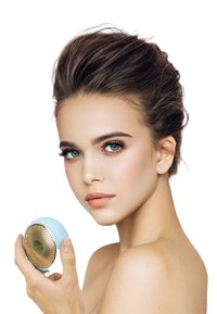 Ljusblå hudvårdsapparat med en strukturerad guldhögtalaryta, hålls av en modell med klar hy och en neutral sminklook.