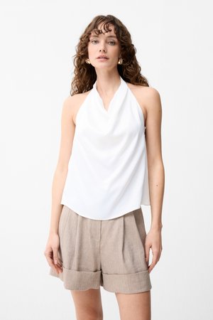 Mujer con cabello rizado que lleva un top halter blanco y pantalones cortos beige de talle alto con dobladillos remangados, de pie contra un fondo liso.