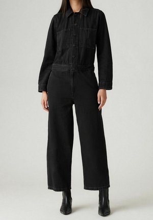 Kobieta ubrana w czarny dżinsowy kombinezon z długimi rękawami, zapinany na guziki z przodu, z kieszeniami na piersiach, szerokimi nogawkami i czarnymi botkami, stojąca na jednolitym tle.