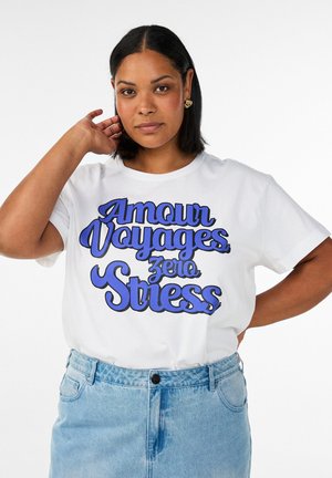 Femme à la peau mi-foncée et aux cheveux noirs portant des boucles d'oreilles en or, un t-shirt blanc avec un texte bleu « Amour Voyages zero Stress » et un jean bleu clair.