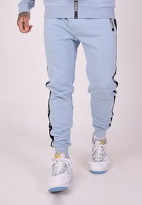 Lichtblauwe sweatpants met zwarte zijkanten met tekst, aangesloten boorden en een zachte textuur, gecombineerd met witte sneakers.