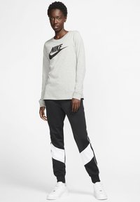 Maglietta grigia chiara a maniche lunghe della Nike con logo nero, abbinata a pantaloni joggers neri con dettagli bianchi e sneakers bianche, in uno sfondo neutro.