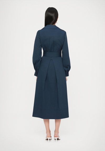 Granatowa sukienka z długim rękawem, dopasowana w talii, z zakładką z tyłu i paskiem do wiązania. Gładka tkanina i fason sięgający do ziemi.